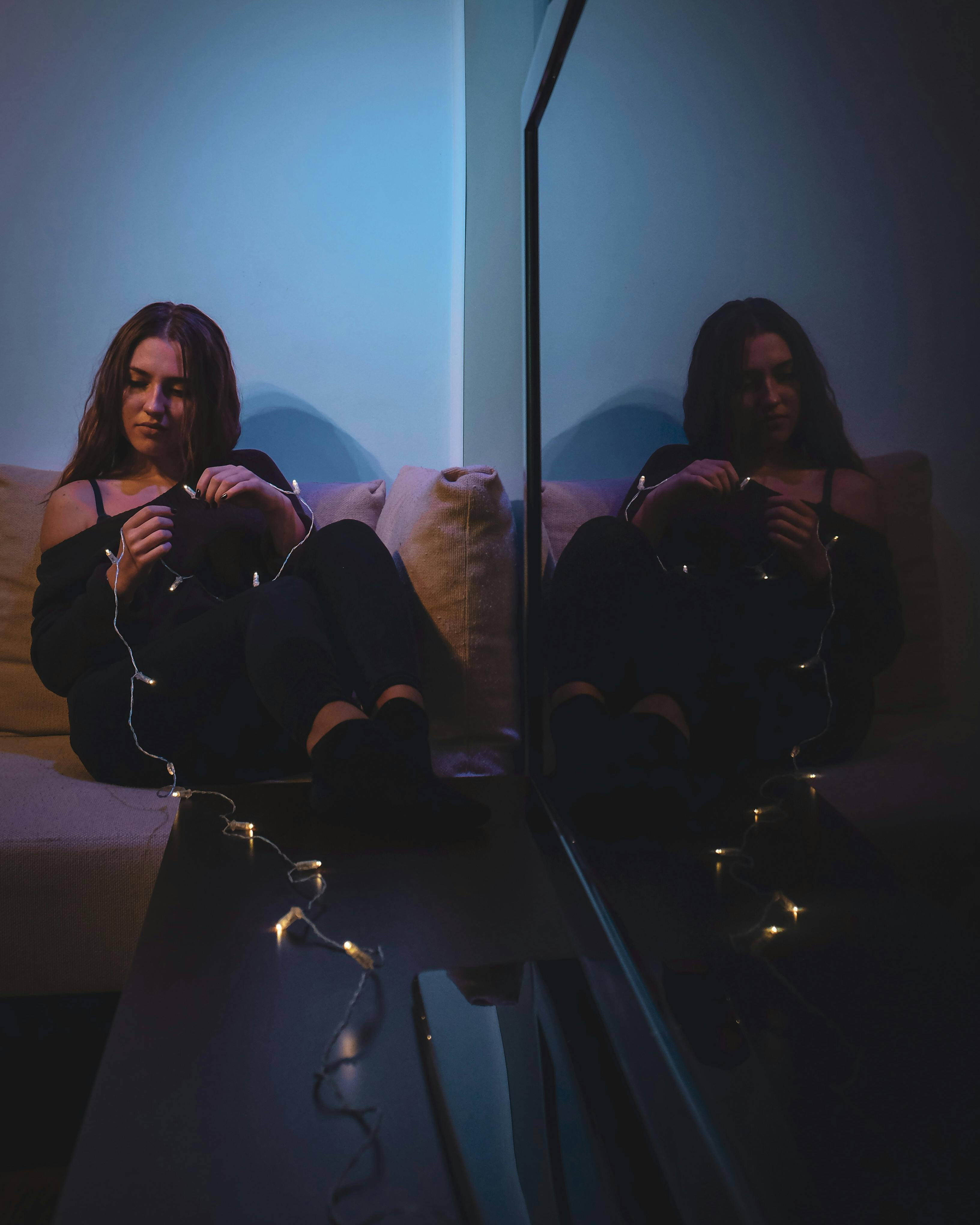 A young woman sitting indoors, reflected in a mirror, holding string lights in a dark, moody atmosphere.