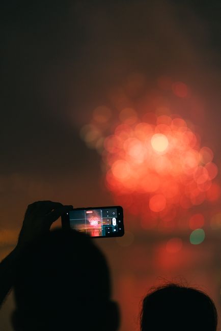 Silhouette of a person capturing vibrant fireworks with a smartphone at night.