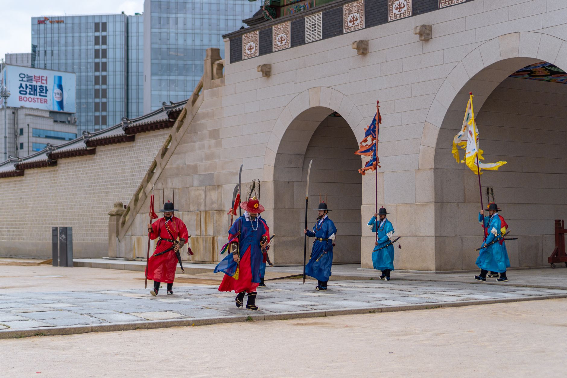 Traditional guards in colorful attire perform a ceremonial walk at a palace gate in a vibrant city setting.