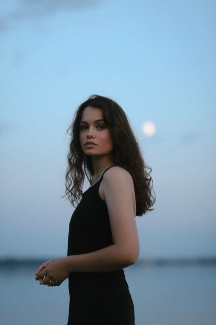 A young woman stands by the sea at twilight, with the moon in the background, capturing serene beauty.