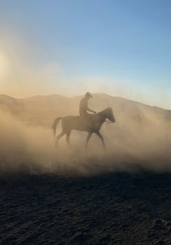 A cowboy riding a horse through dust with mountains in the background during sunset.