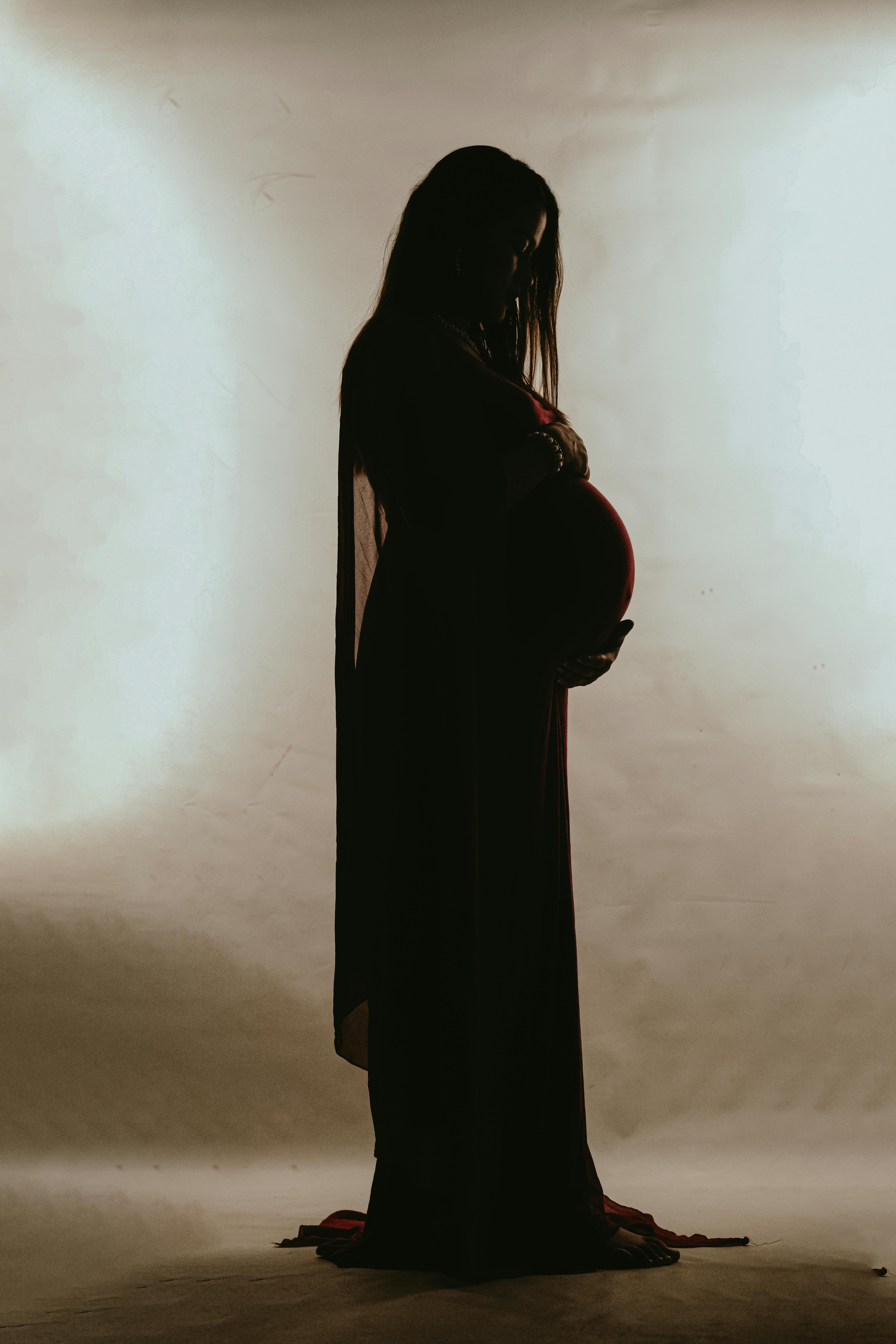 Artistic silhouette of a pregnant woman in a dress posing gracefully in a studio setting.