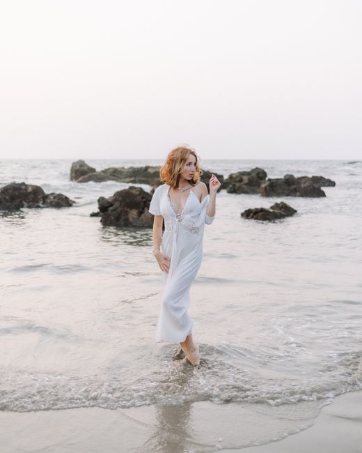 A woman in a white dress elegantly poses on the rocky beach shore at sunset.