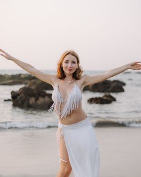 Smiling woman in white bikini and skirt at a serene beach during sunset.