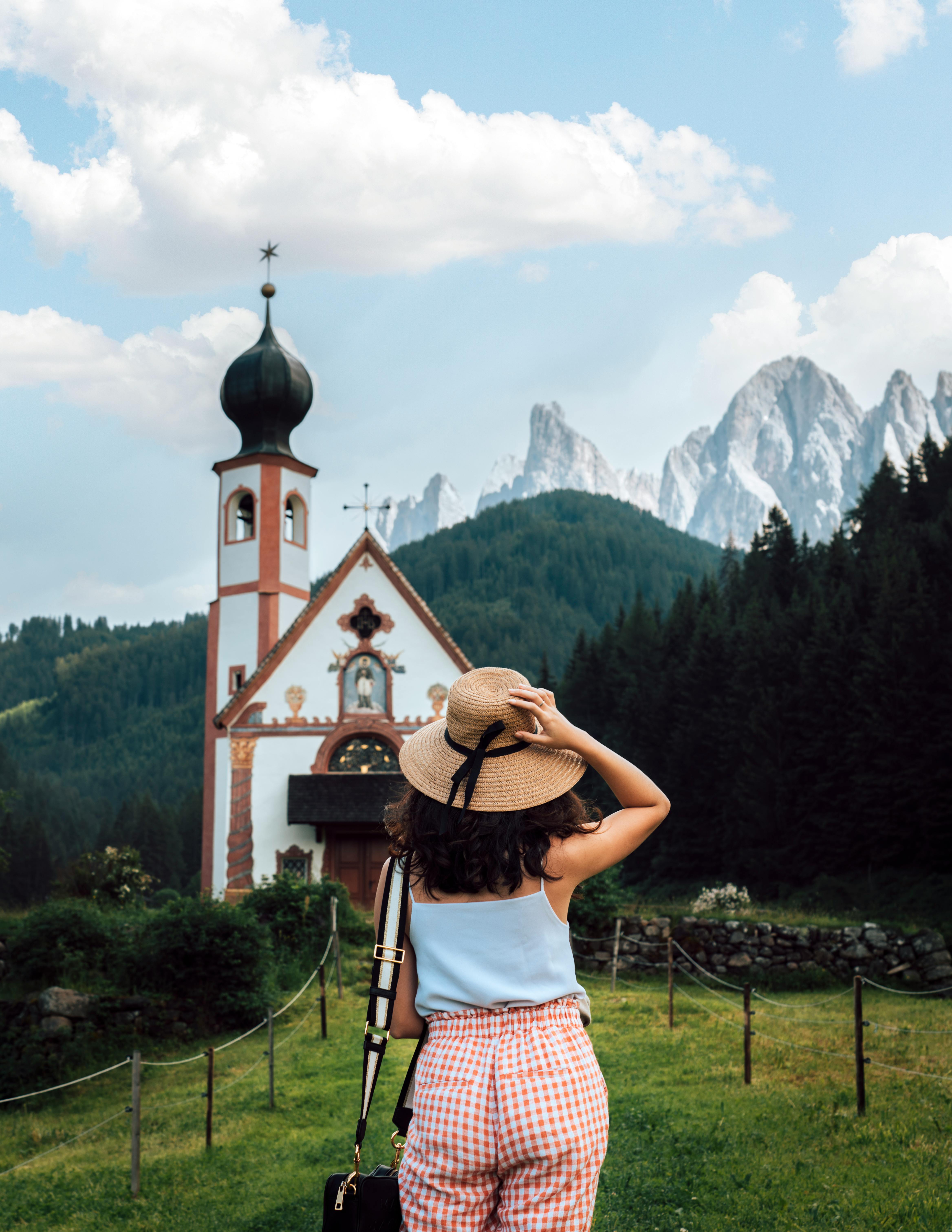 Woman in a hat gazing at the scenic church and mountain landscape of St. Magdalena, Italy, showcasing tranquility and beauty.