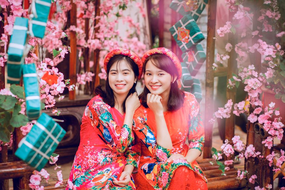 Two smiling Asian women in floral dresses sitting amidst cherry blossoms and festive decorations, celebrating with joy.