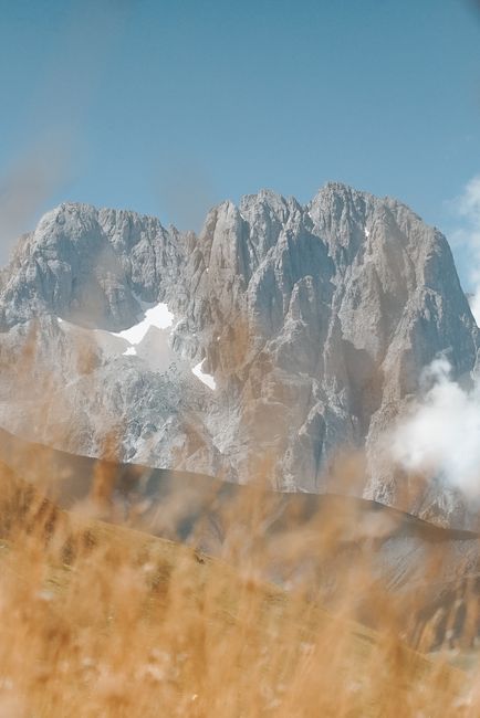 A breathtaking view of a rocky mountain peak surrounded by tall grass under a clear blue sky.