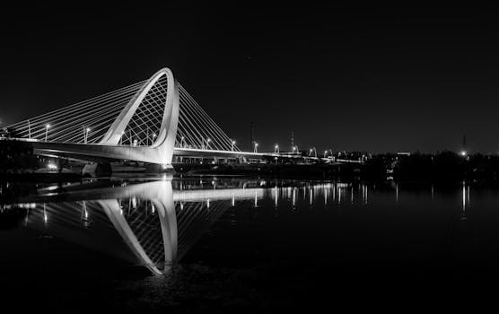 A captivating black and white photo of an illuminated bridge with perfect water reflections at night.