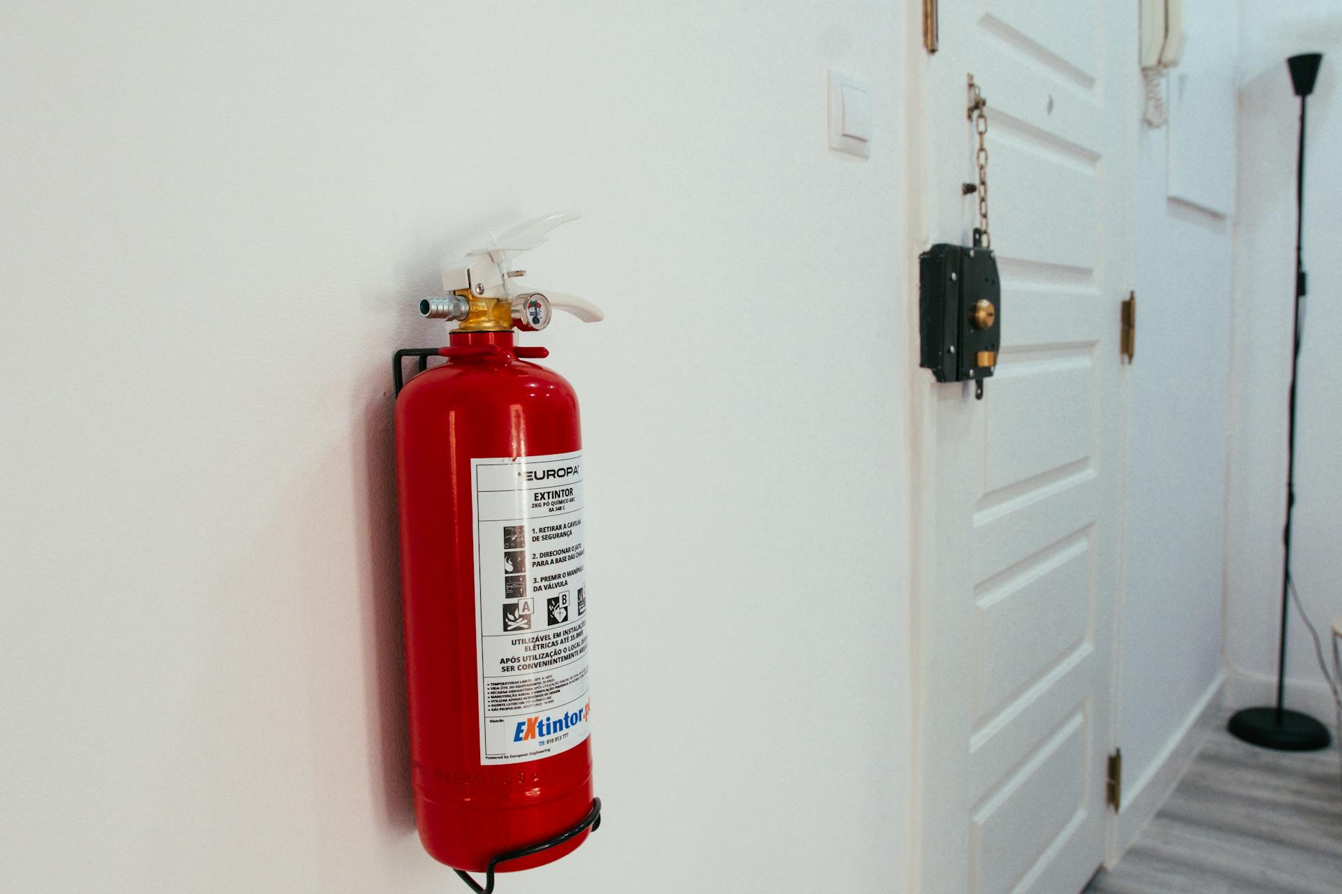 Red fire extinguisher mounted on a white wall in a modern apartment hallway.