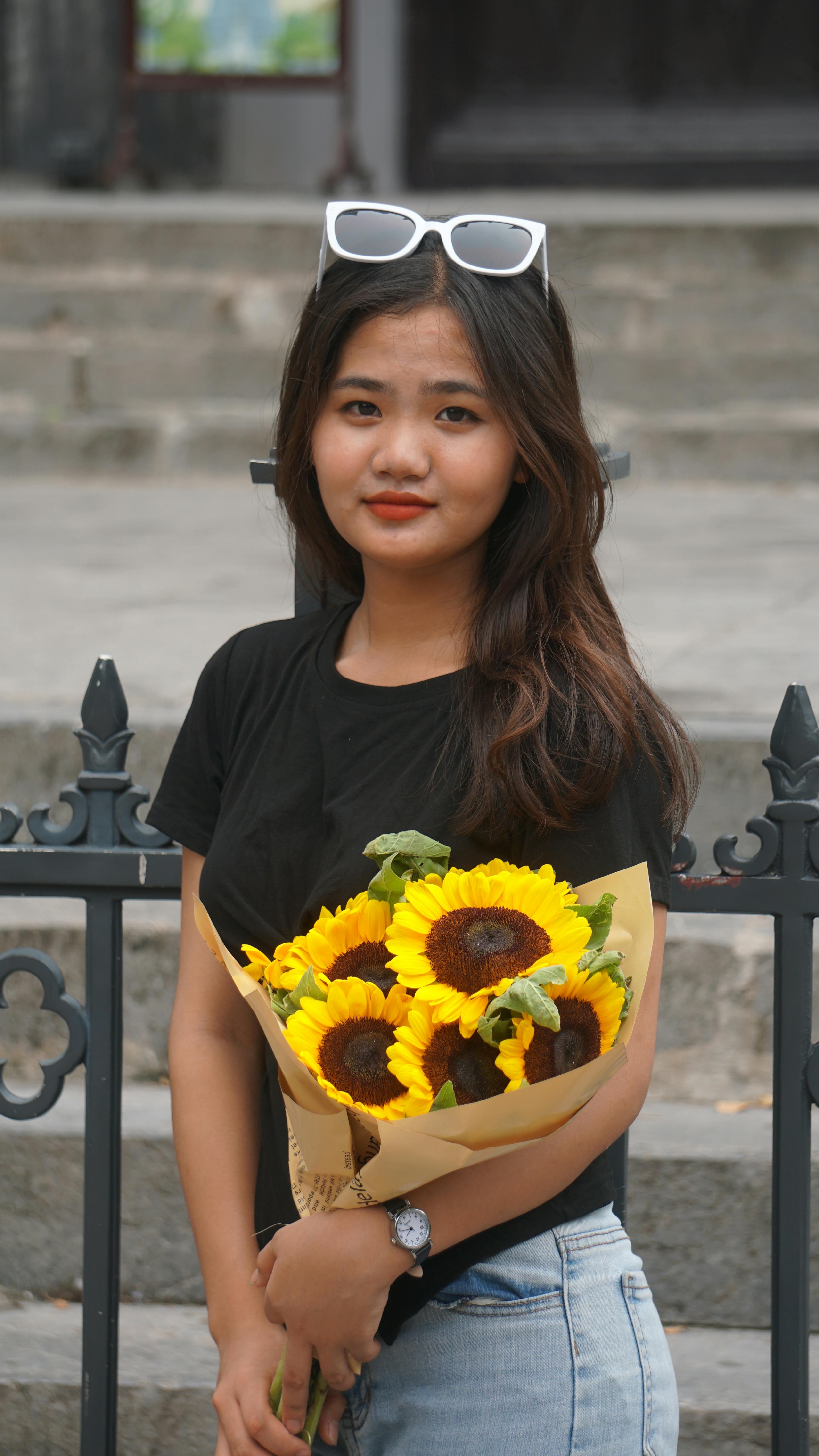 A young woman stands outdoors holding a vibrant bouquet of sunflowers, exuding a stylish and summery vibe.