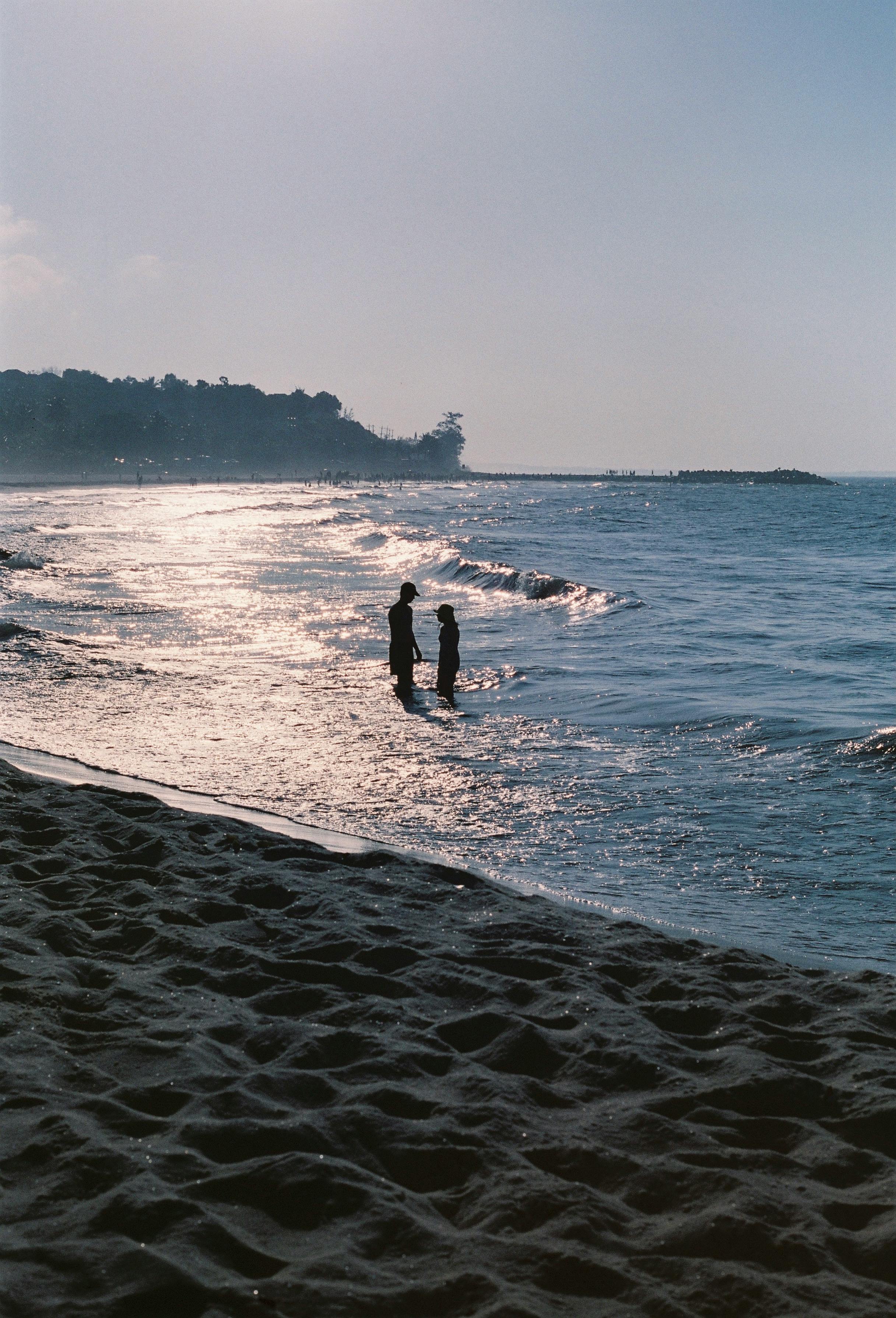 Silhouetted figures stand by the water on a calm beach during sunset.
