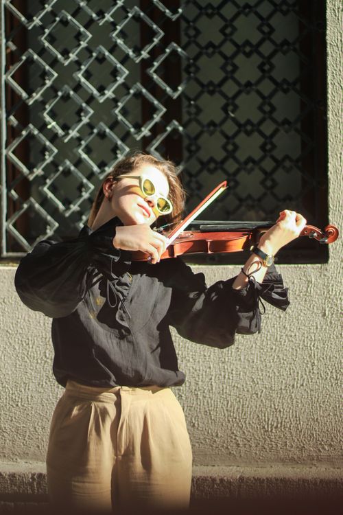 Free Young woman plays violin outdoors in sunlight, wearing sunglasses, against a decorative window backdrop. Stock Photo