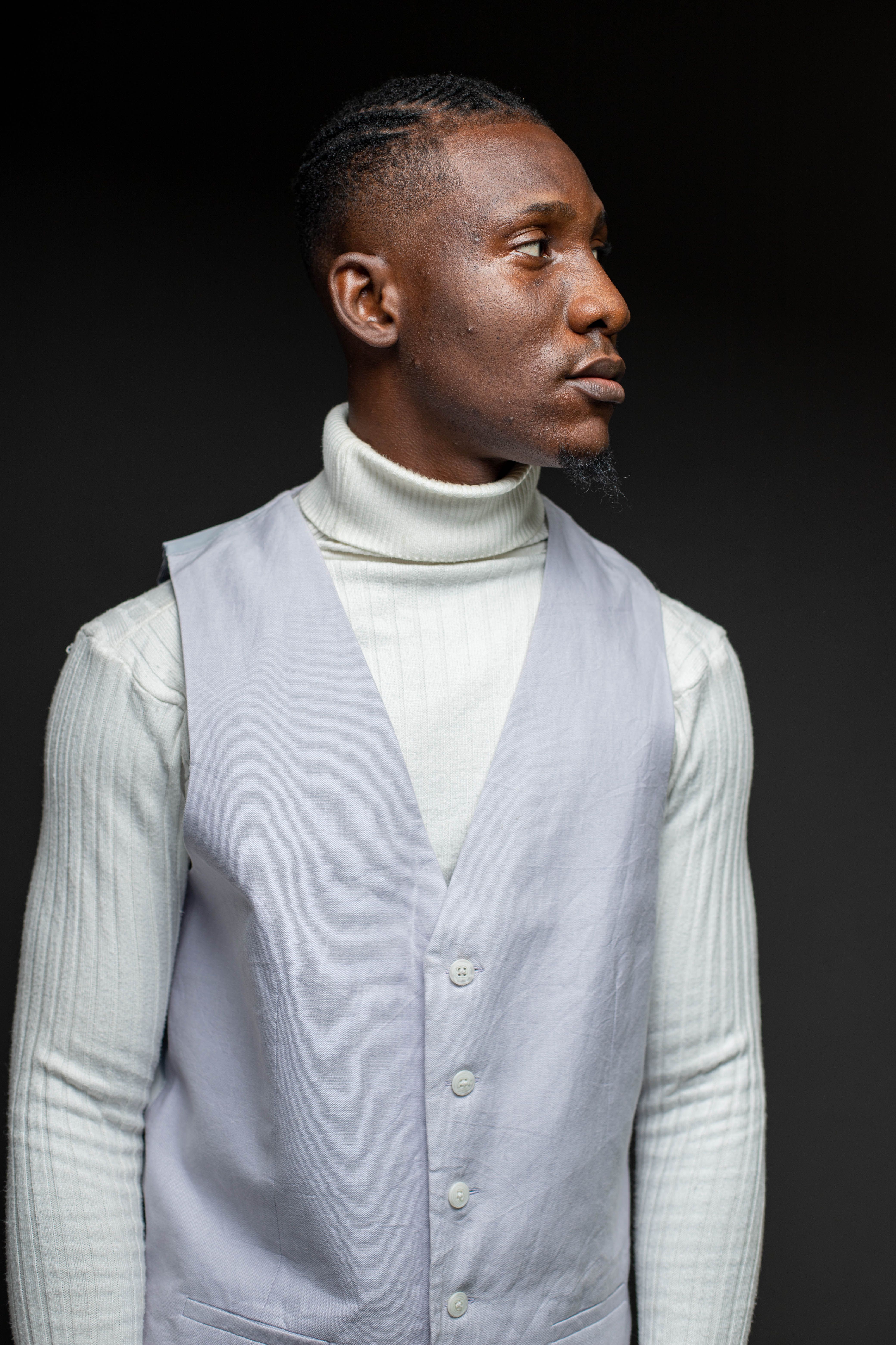 A fashionable young man poses in a studio wearing a turtleneck and vest, showcasing modern elegance.