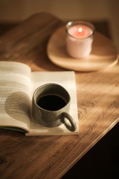 A warm setting with a coffee cup, open book, and candle on a wooden table in sunlight.