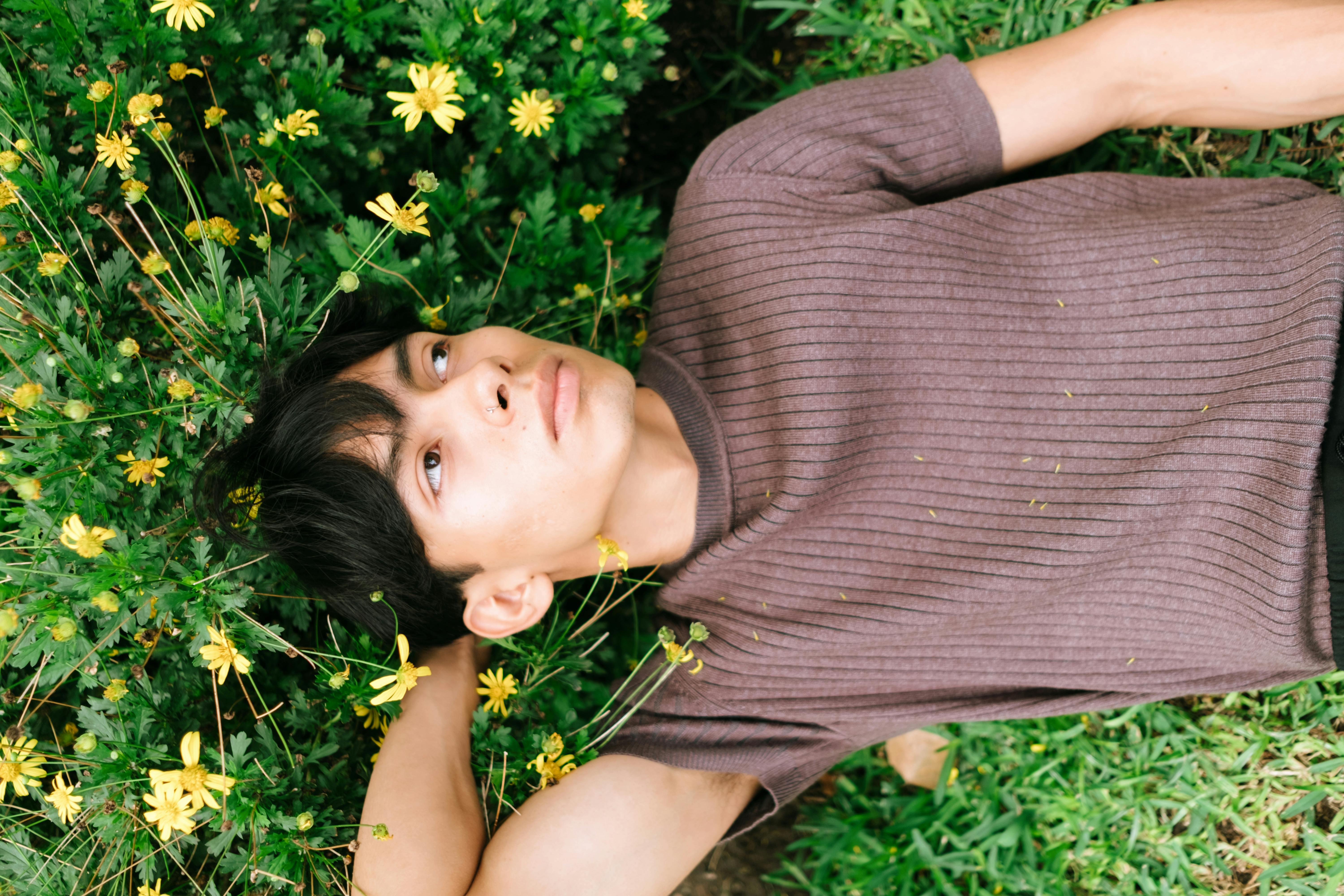 A man in a brown t-shirt lies among yellow flowers in a green meadow, enjoying a relaxed moment outdoors.
