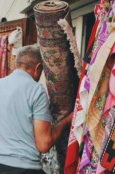 Man holding a traditional carpet amid vibrant textiles in an outdoor market setting.