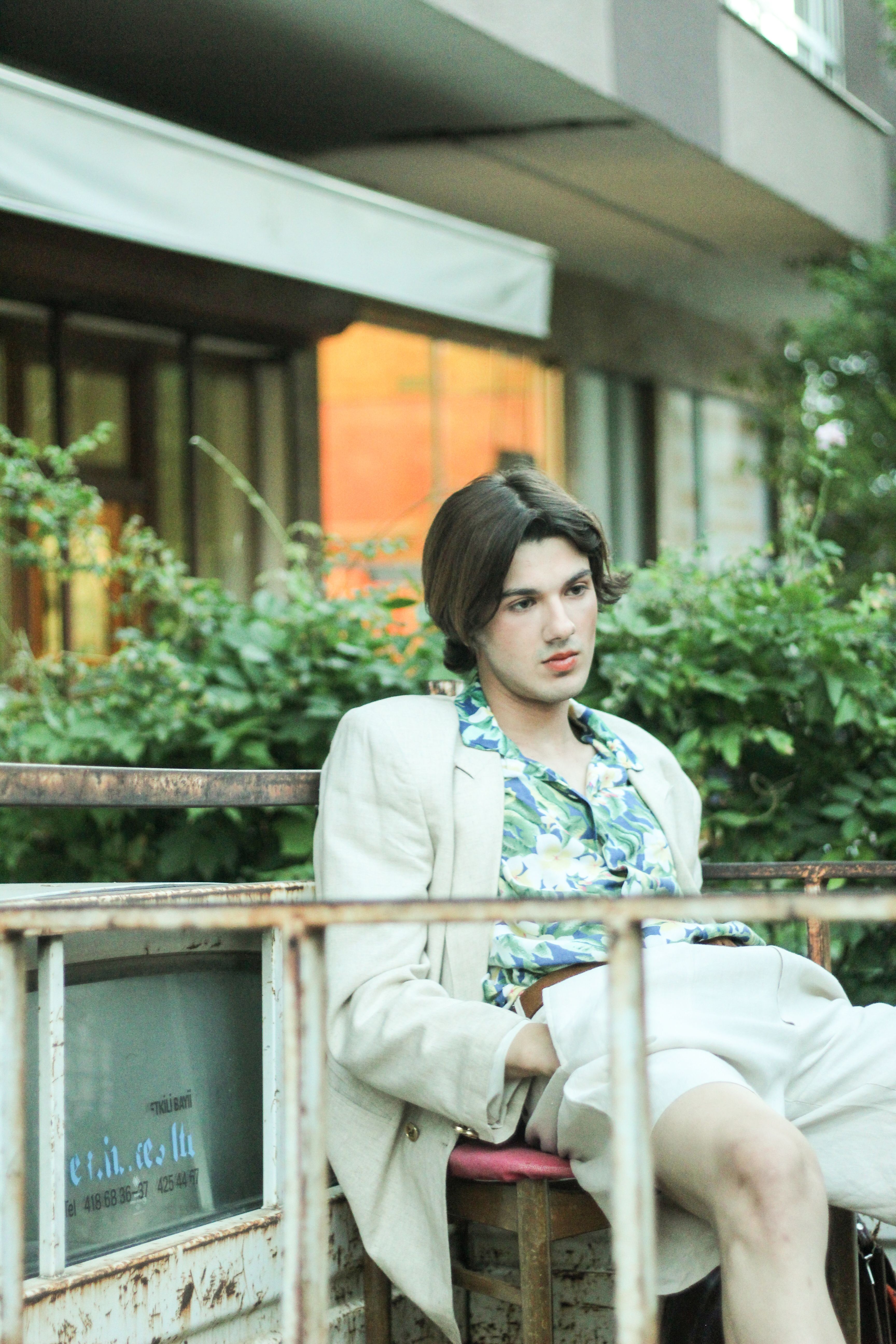 A stylish man in a floral shirt and linen suit sits outdoors, exuding relaxation.
