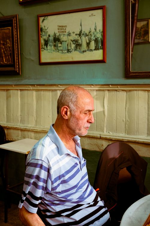 Free Senior man sitting in a cozy café, wearing a striped polo shirt, bathed in warm light. Stock Photo