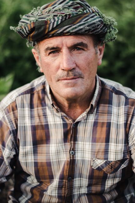 Middle-aged man wearing a checkered shirt and traditional headscarf in an outdoor setting.