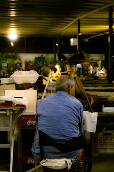 Back view of a man sitting at a bustling night market stall surrounded by wares and people.