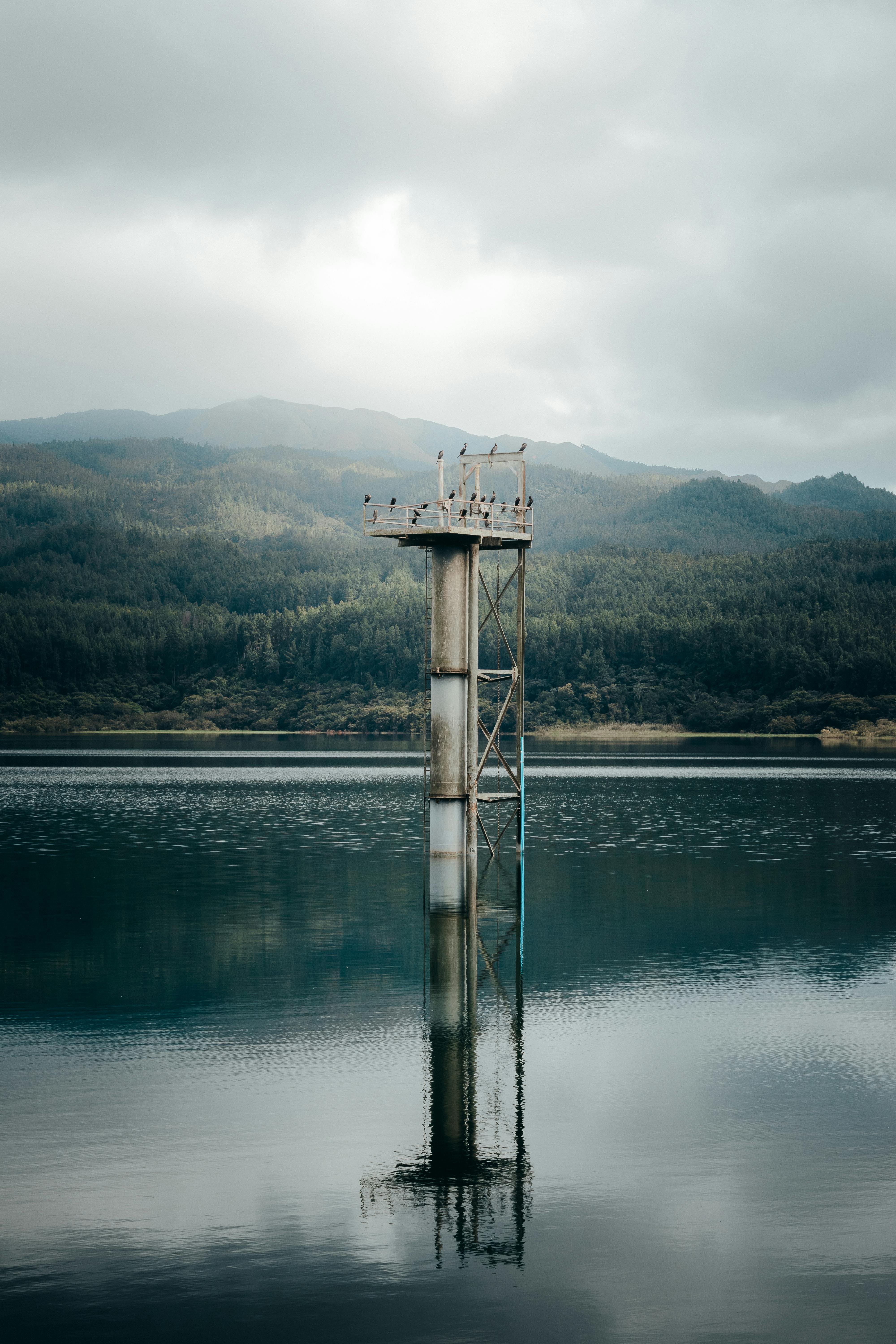 Solitary tower stands amidst calm lake waters with mountainous background, under an overcast sky.