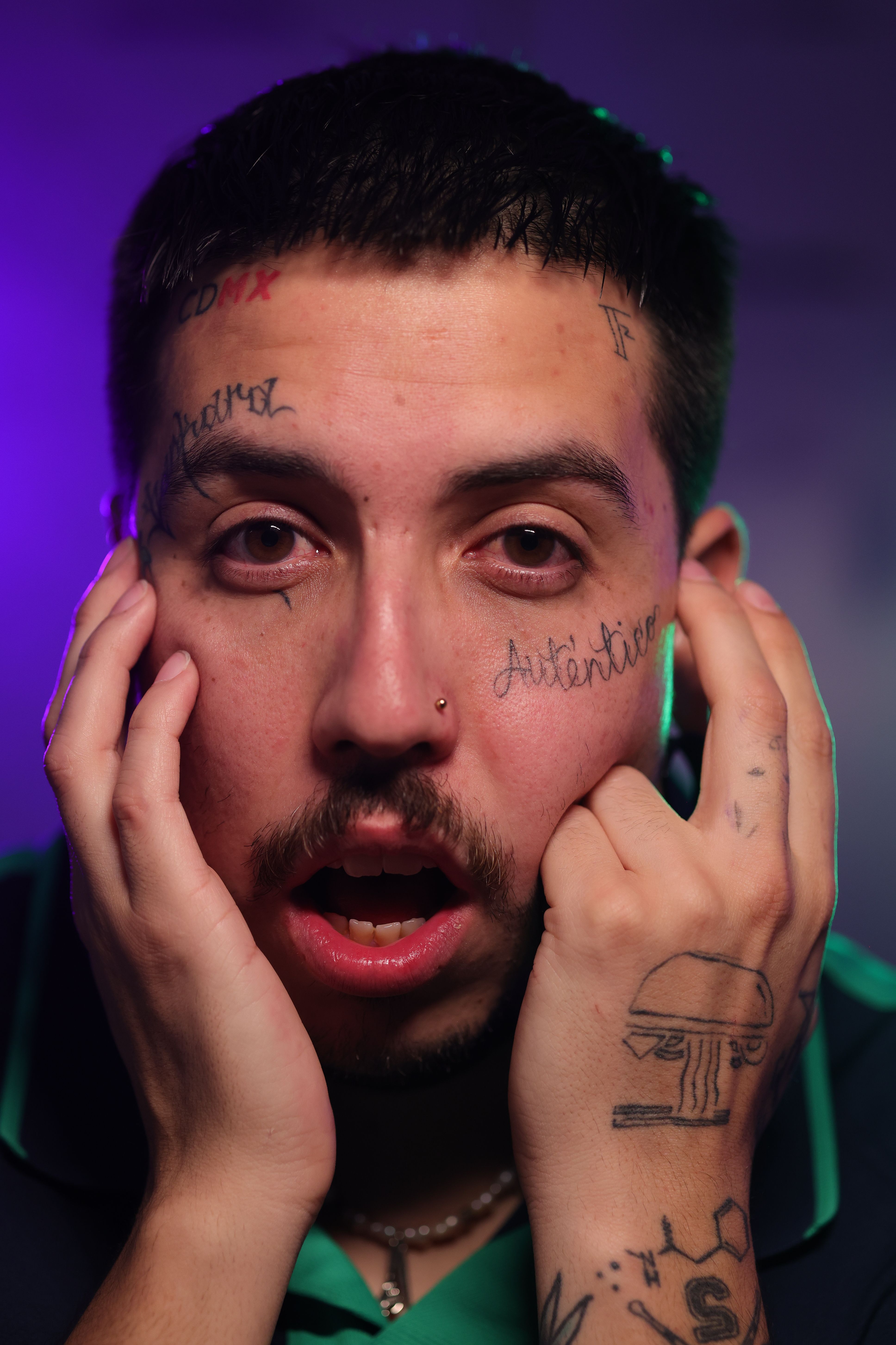 Close-up of a tattooed man with an expressive open mouth and hands on face.