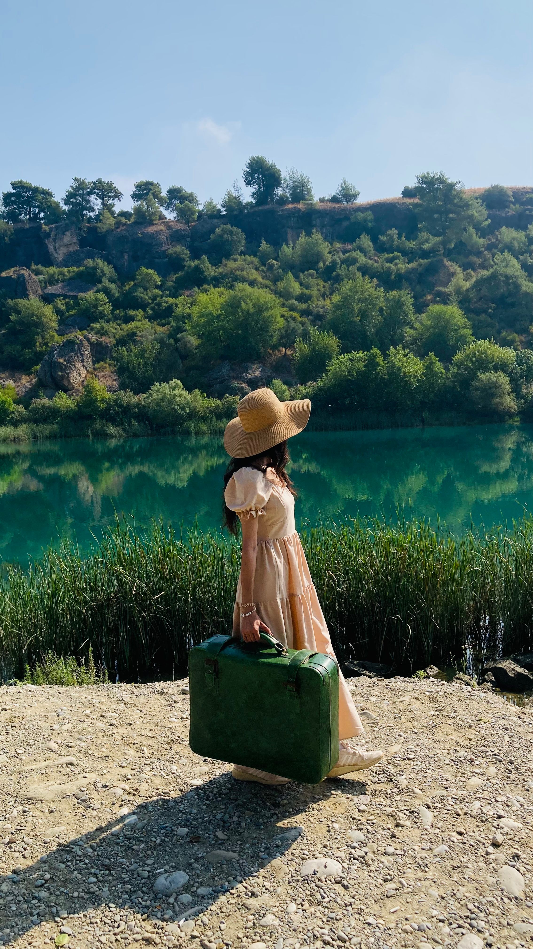 A woman in a dress and hat carries a green suitcase by a tranquil river in a summer landscape.
