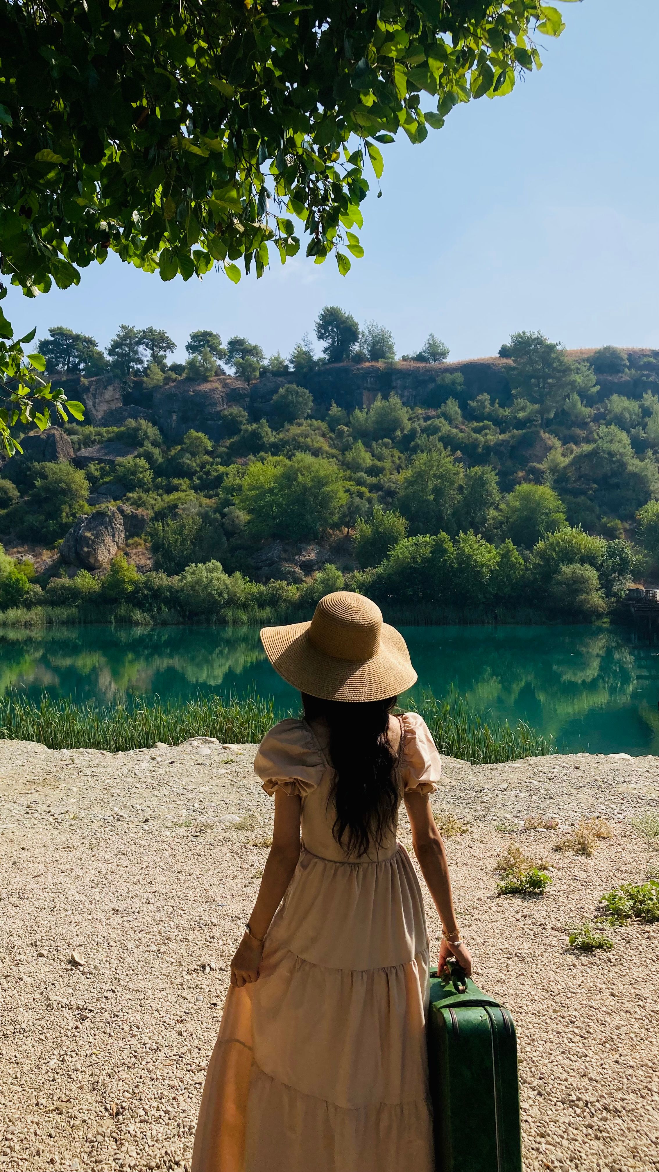 A woman in a sundress and hat stands by a tranquil lake holding a suitcase.