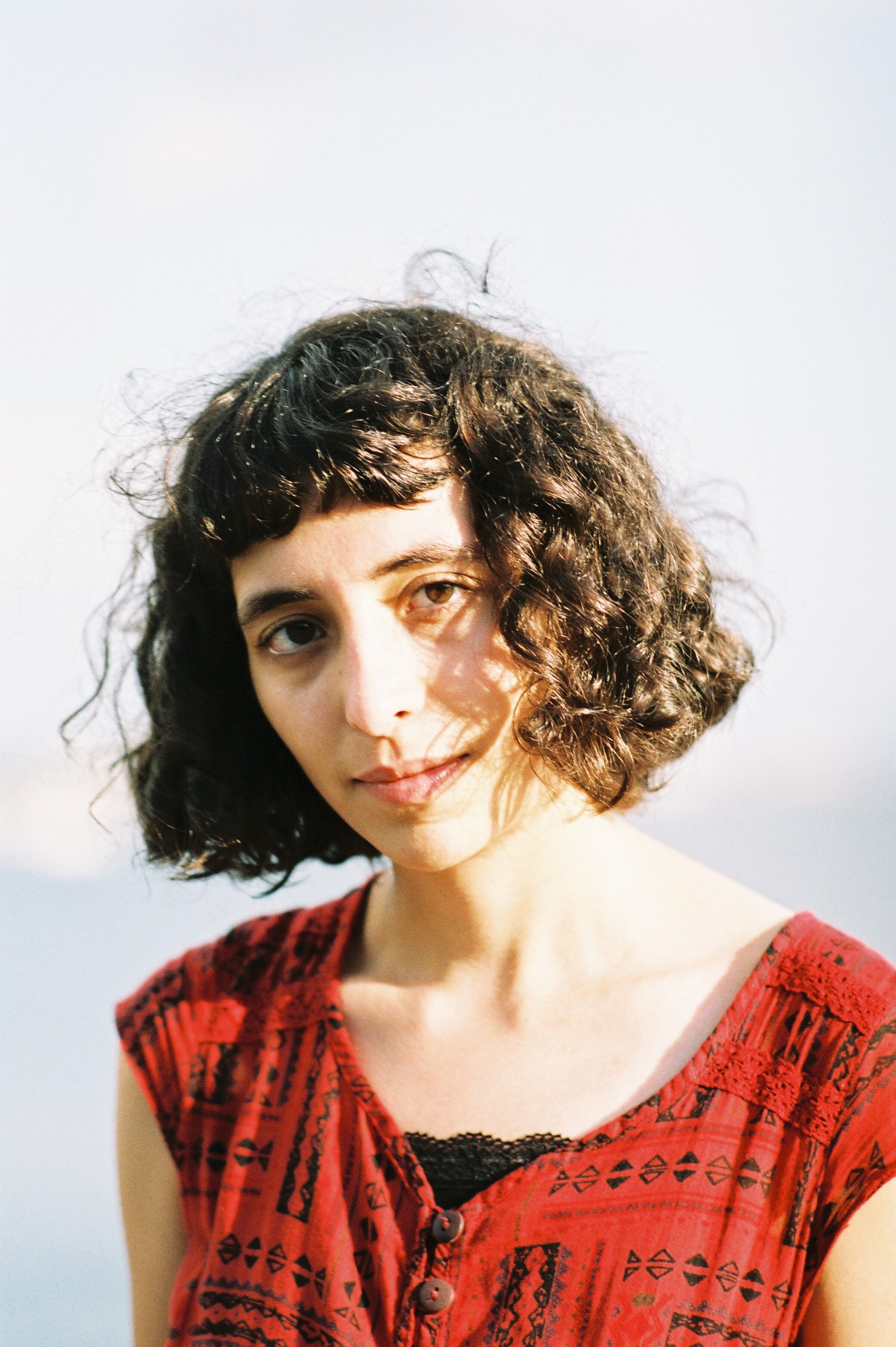 A serene portrait of a woman with curly hair wearing a red dress, outdoors with natural lighting.