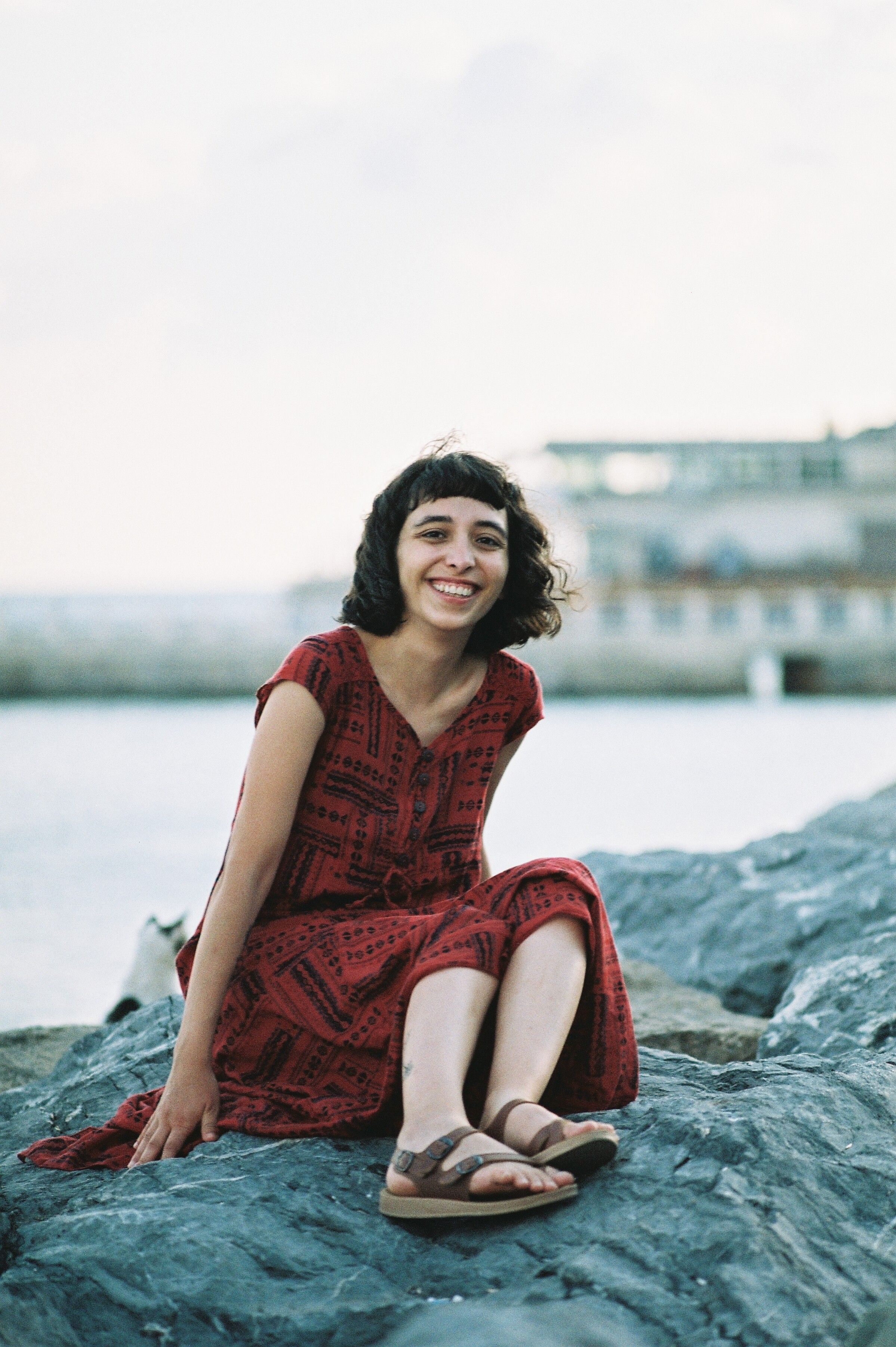 Young woman in red dress smiling while sitting on coastal rocks, enjoying a sunny summer day.