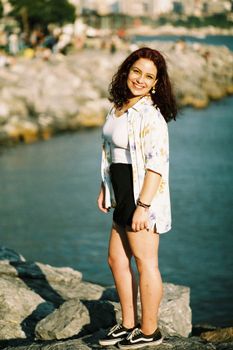 A young woman with curly hair smiling, standing on a rocky shore by the ocean on a summer day.