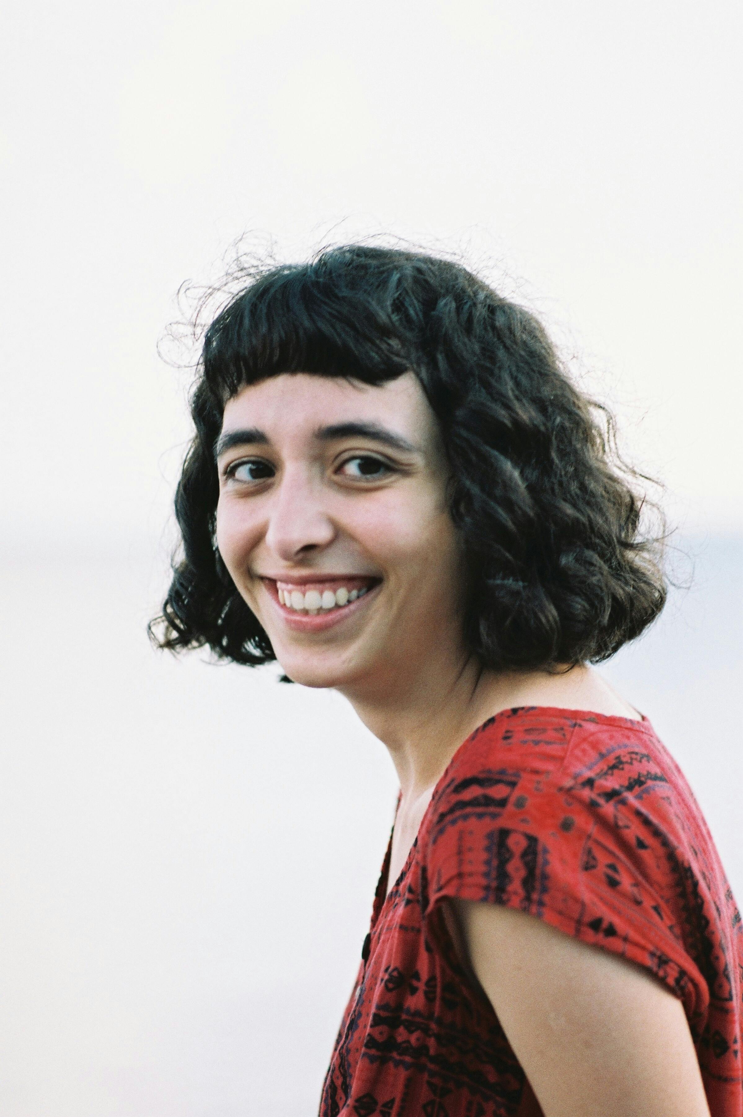 Portrait of a smiling woman with curly hair outdoors wearing a red patterned dress on a bright day.