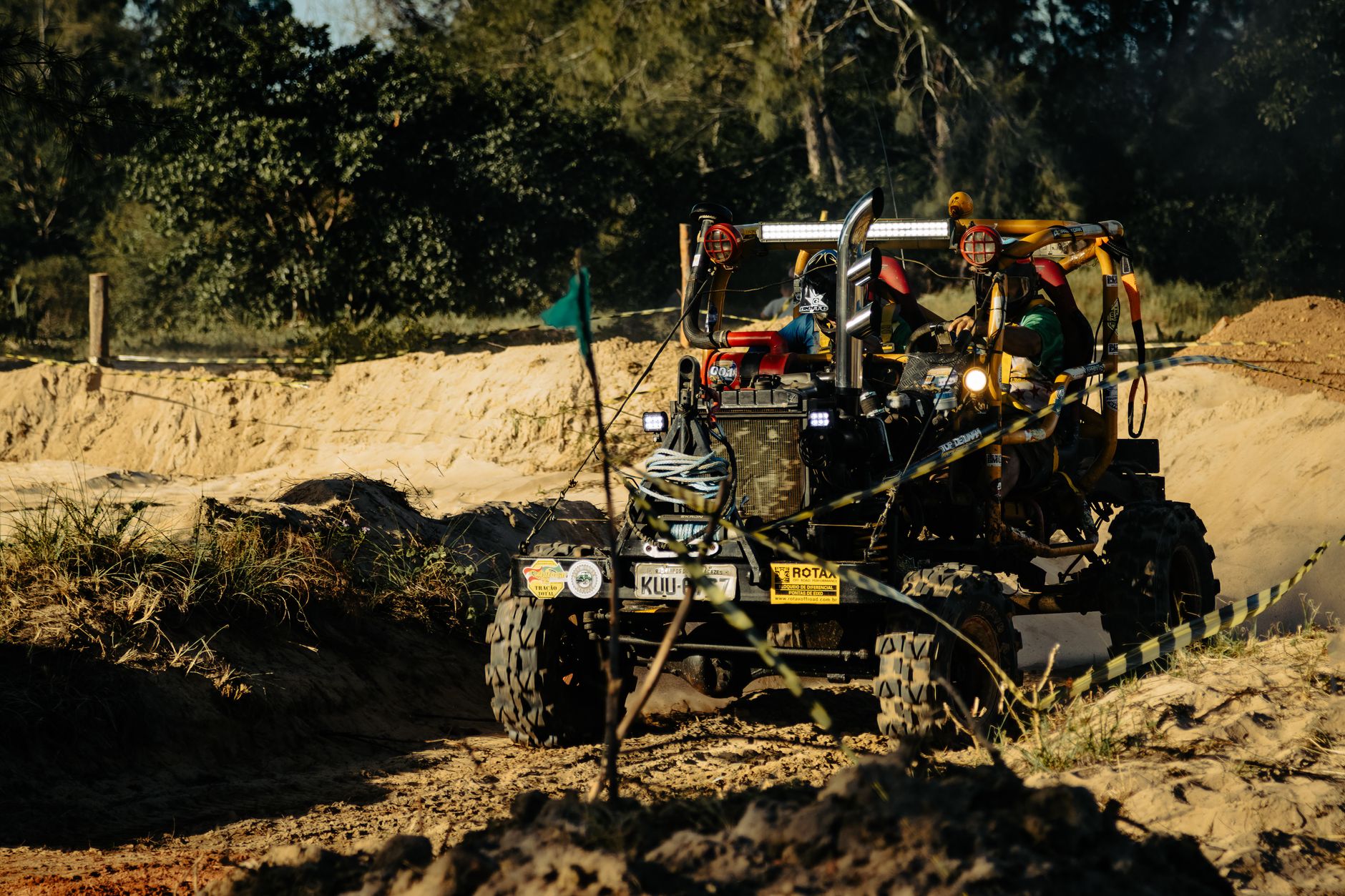 Two drivers in a rugged vehicle navigate challenging off-road terrain in a forest setting.