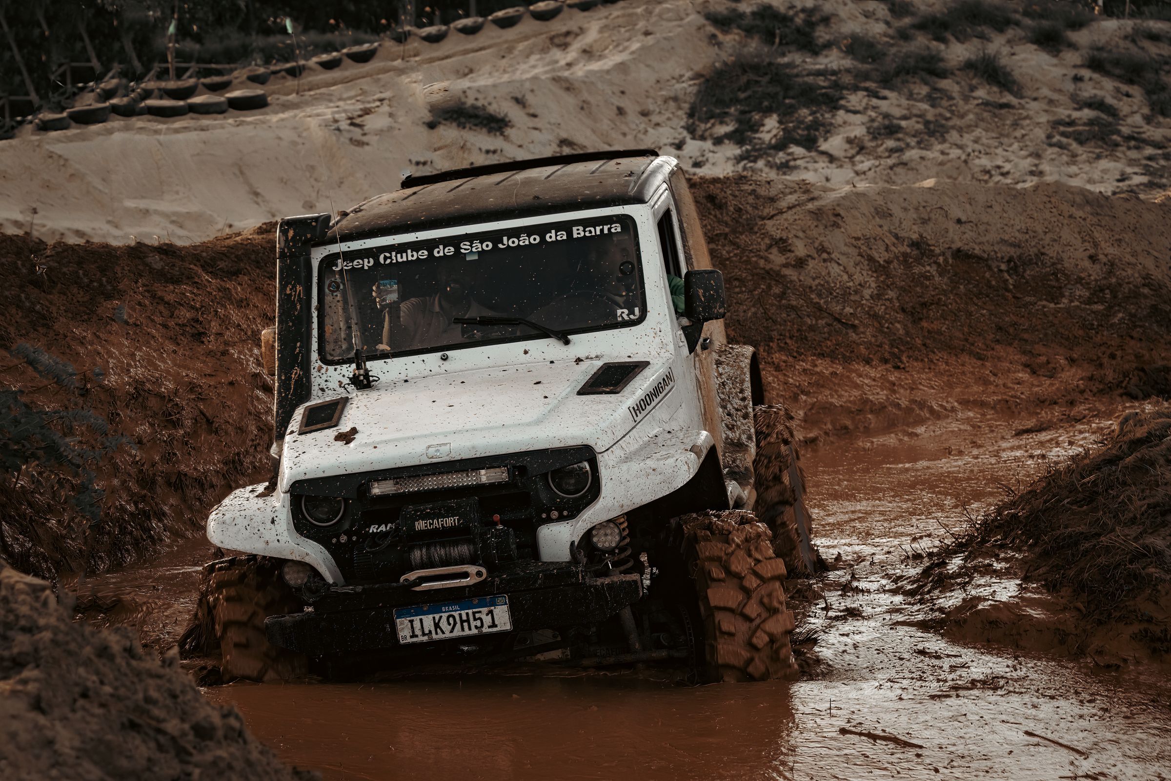 Modified white 4x4 with a front winch crawling through a deep red-mud rut on an off-road course