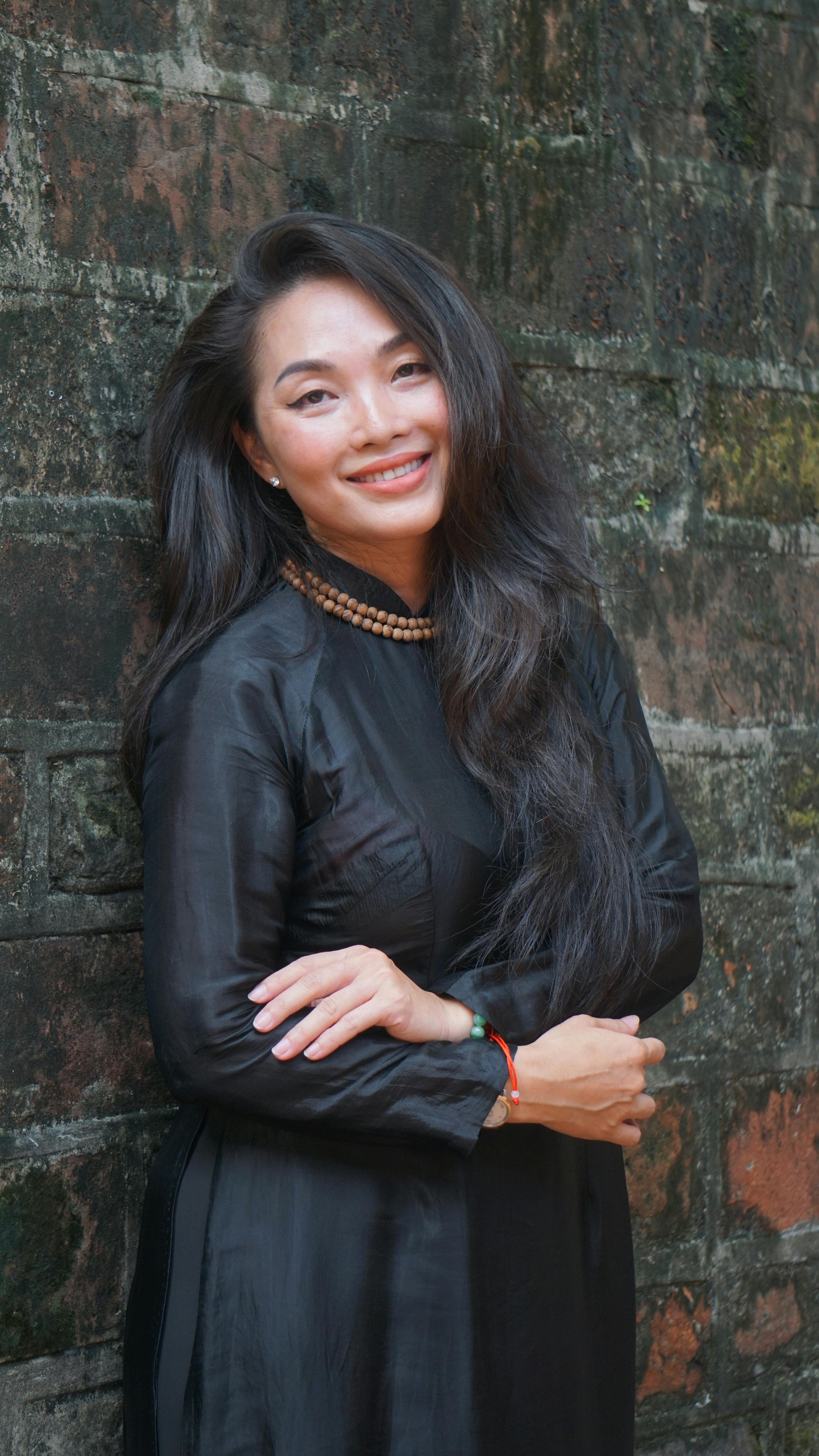 Elegant portrait of a smiling woman with long black hair in a black dress, posing against a textured wall.