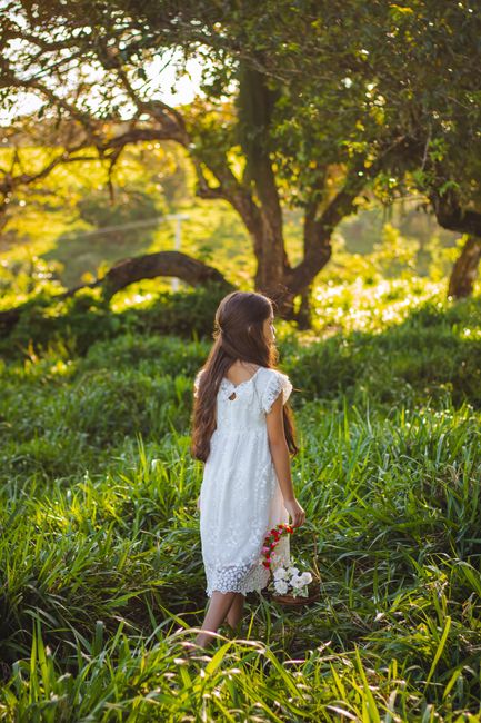 Child with long brown hair holding a flower basket walks in a sunlit meadow.