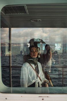 Woman wearing headscarf stands by a ferry window with ocean view reflection.