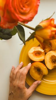 A hand reaches for fresh peach halves on a yellow plate near vibrant roses.