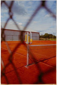 A tennis court with clay surface, net, and scoreboard seen through a fence on a sunny day.