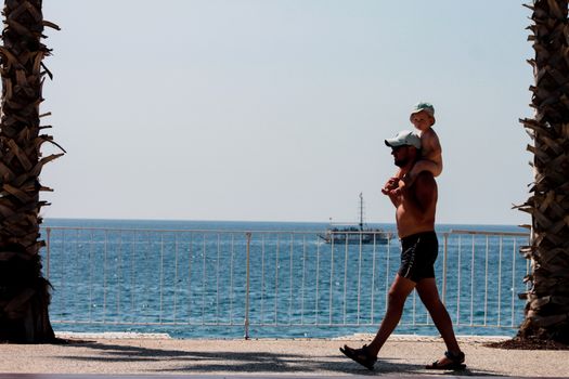 A father carries his child while walking by the seaside promenade, enjoying a sunny vacation day.