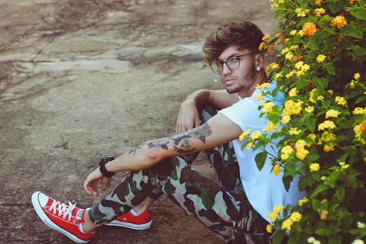 一个时尚的年轻人戴着眼镜、纹着身，坐在户外鲜艳的黄色花朵旁
