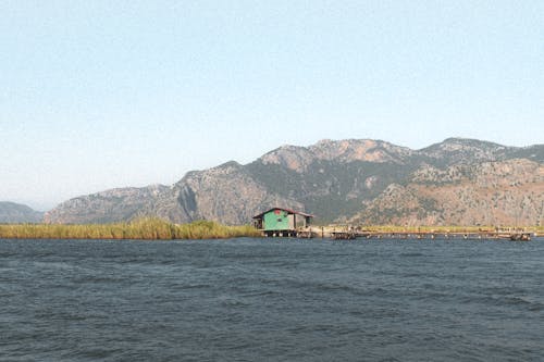 View of a Wood Hut on a Riverbank and Mountains in the Background 