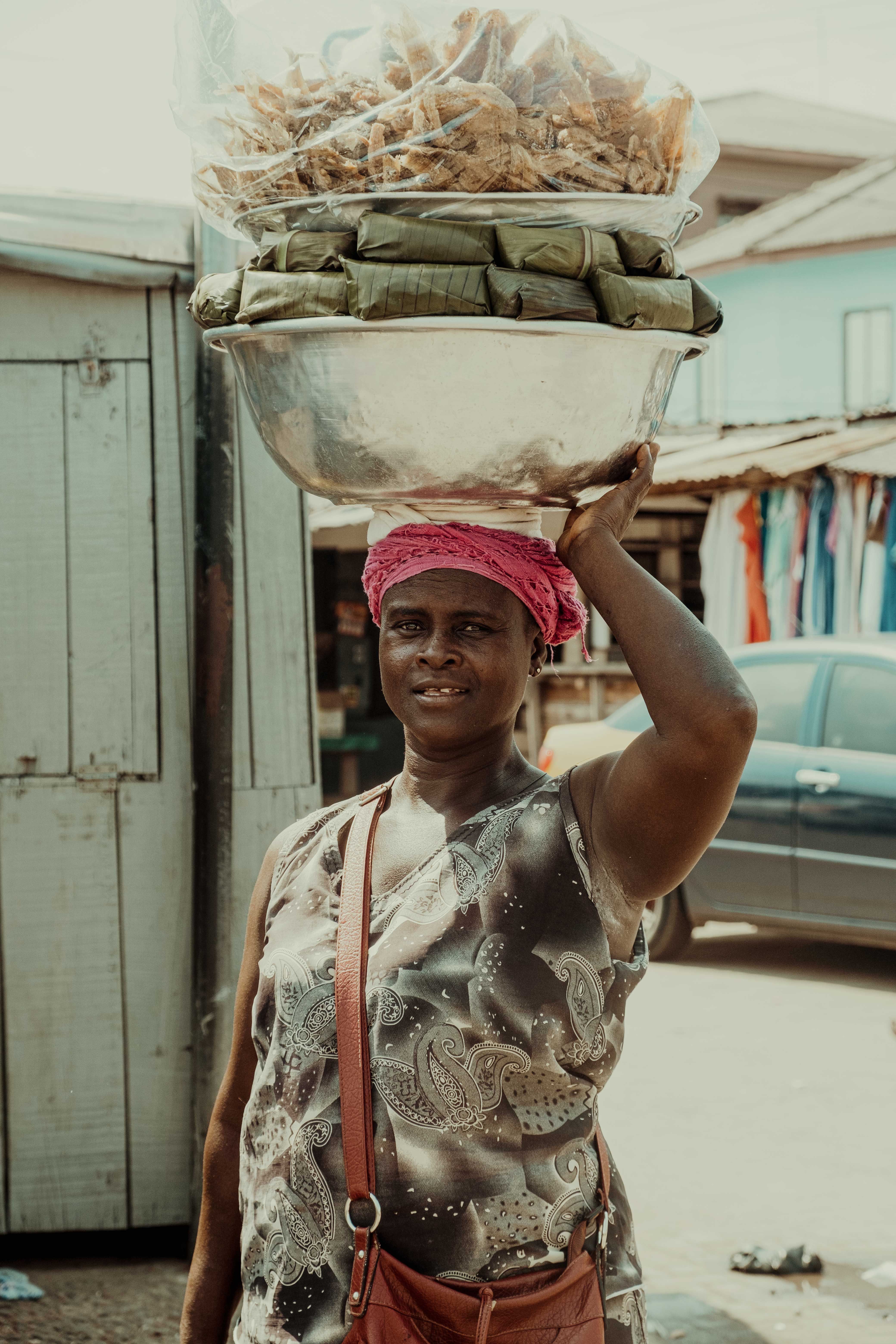 African woman balancing a bowl of goods on her head in a bustling Kumasi street.