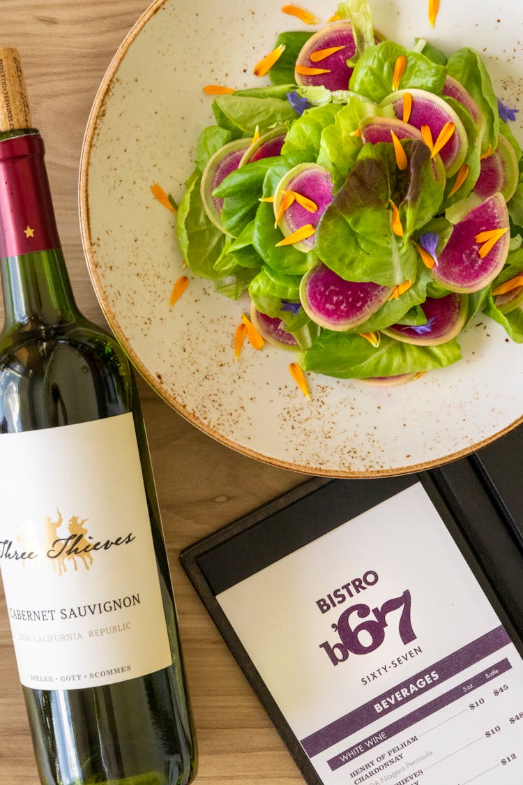 Top View Of A Colorful Salad, A Bottle Of Wine And A Menu Card On A Table In A Restaurant