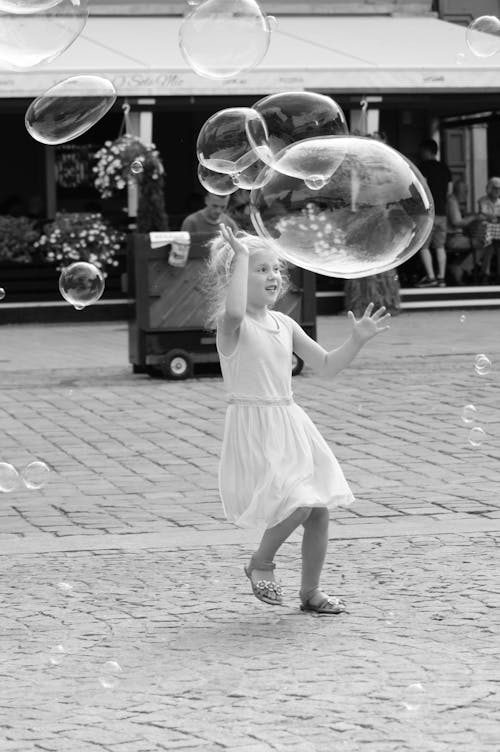 A Little Girl Running among Soap Bubbles on the Pavement 