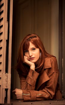 A fashionable redhead woman in a brown jacket poses charmingly by a window indoors.