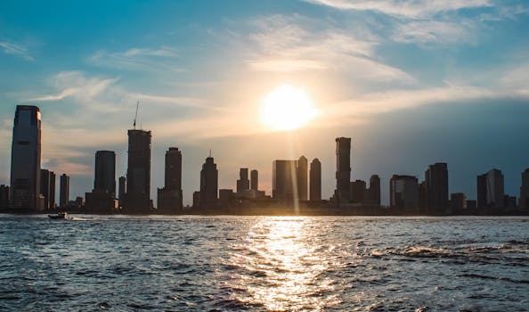 Silhouette of a city skyline at sunset with water reflecting the sunlight, highlighting urban structures.