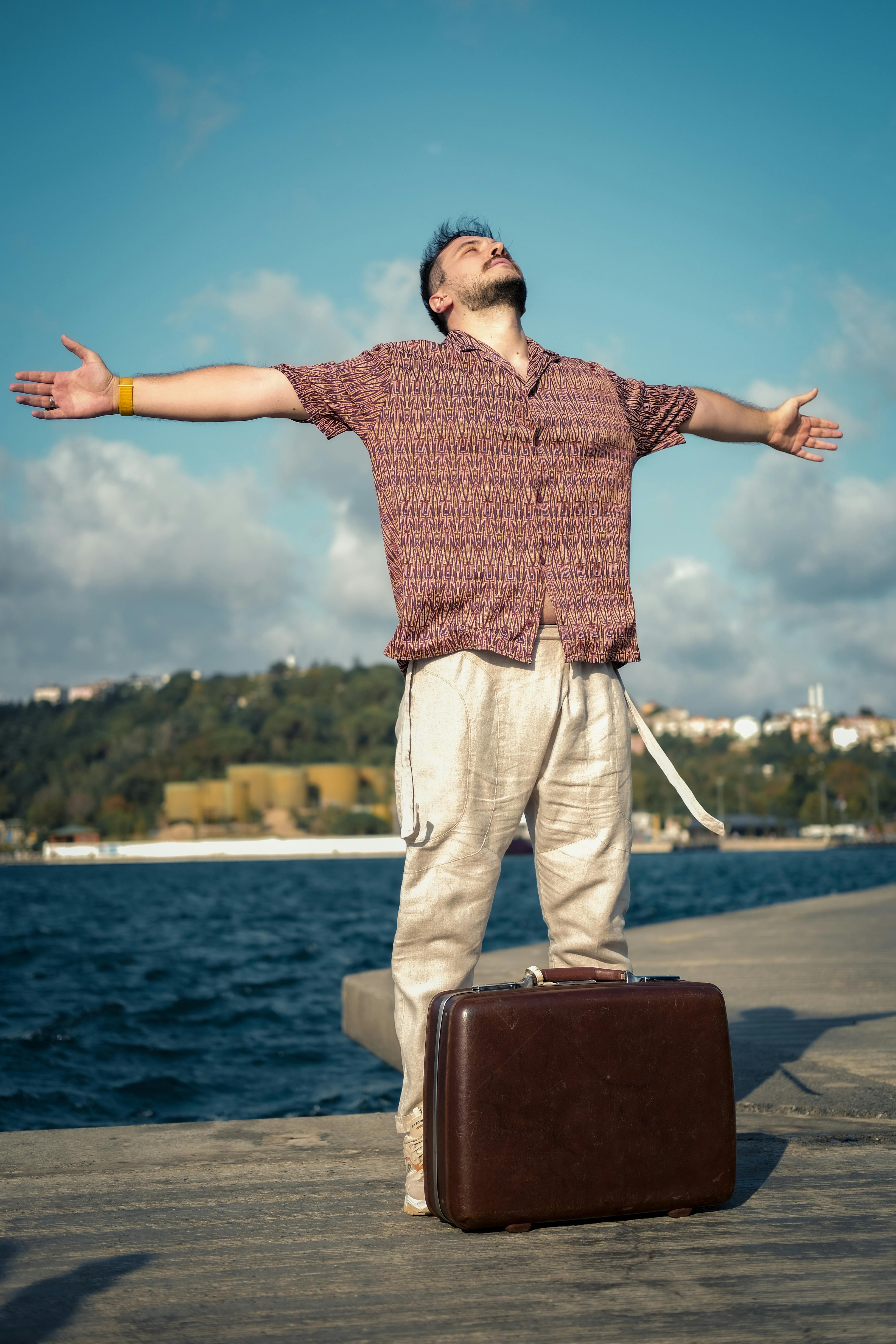 A man with open arms stands by the sea, embracing freedom with a suitcase.