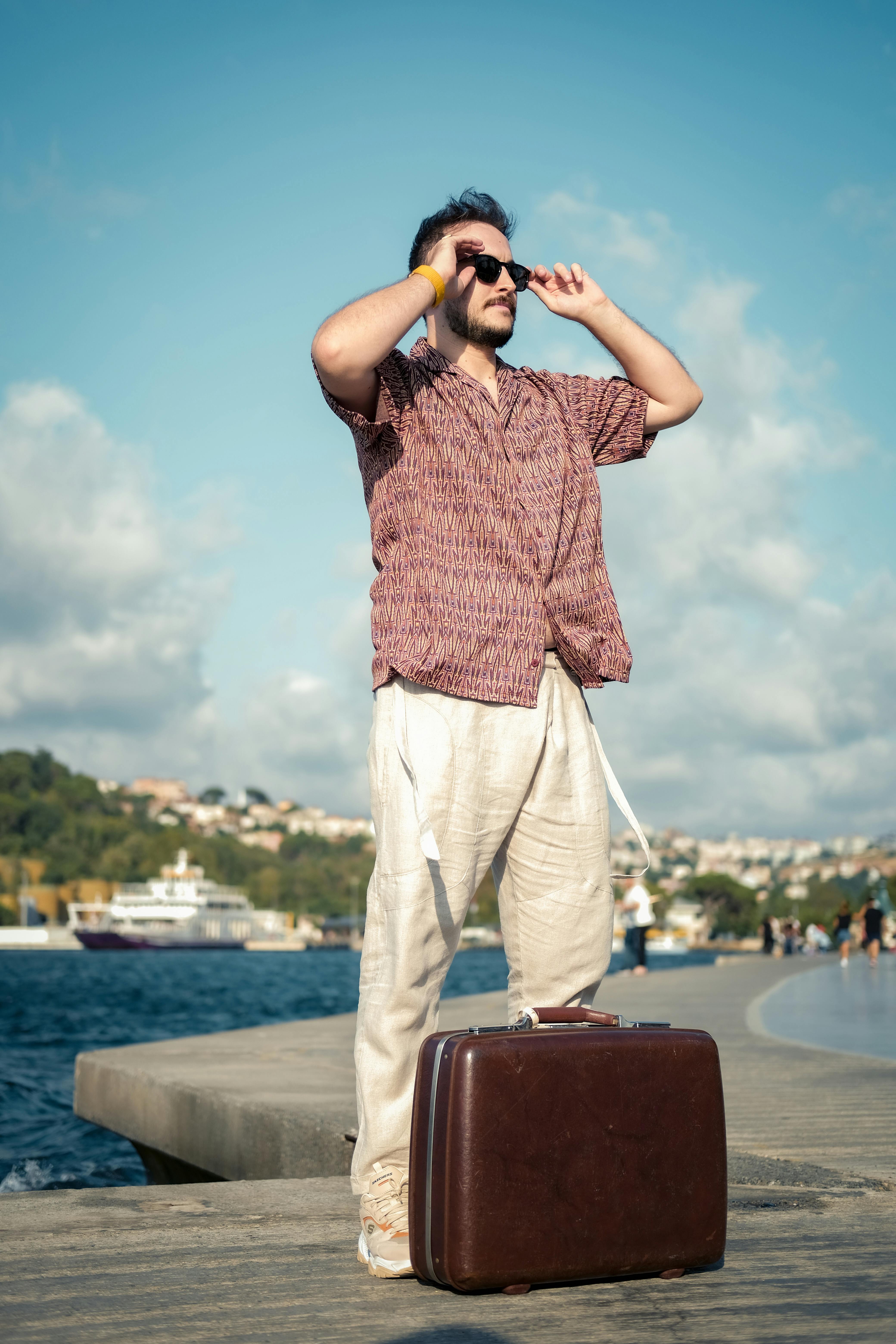 Man standing on seaside promenade adjusting sunglasses, holding a suitcase.