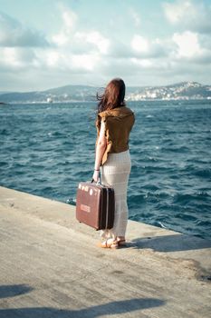 A woman stands on a beach promenade holding a vintage suitcase by the sea.